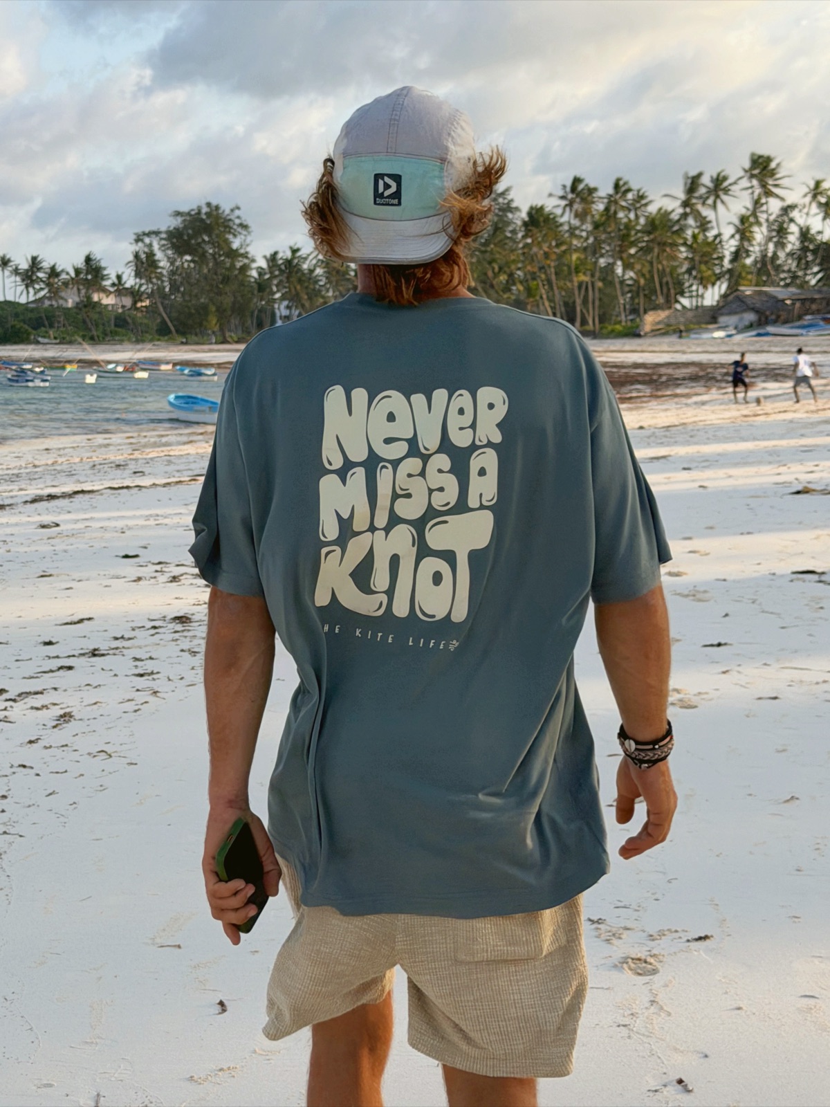 The Teal Blue Never Miss A Knot tee worn on the beach, back print showing