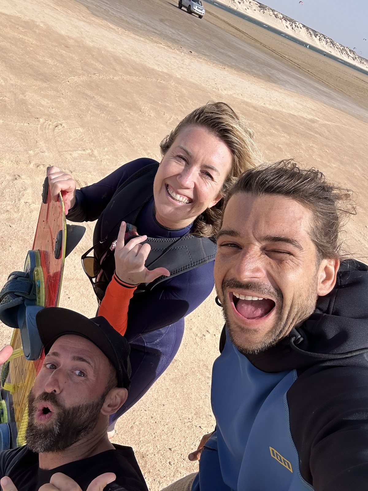 Three riders laughing in a selfie on the Dakhla dune, lagoon behind